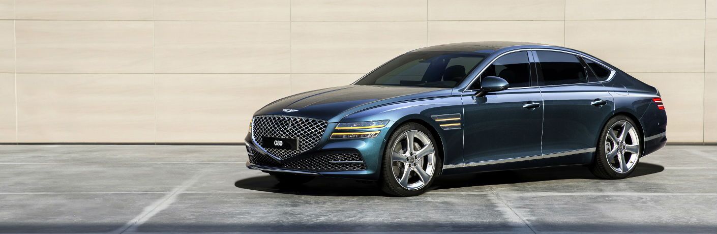 A sleek blue luxury Genesis G80 sedan parked on a smooth concrete surface with a beige modern wall in the background.