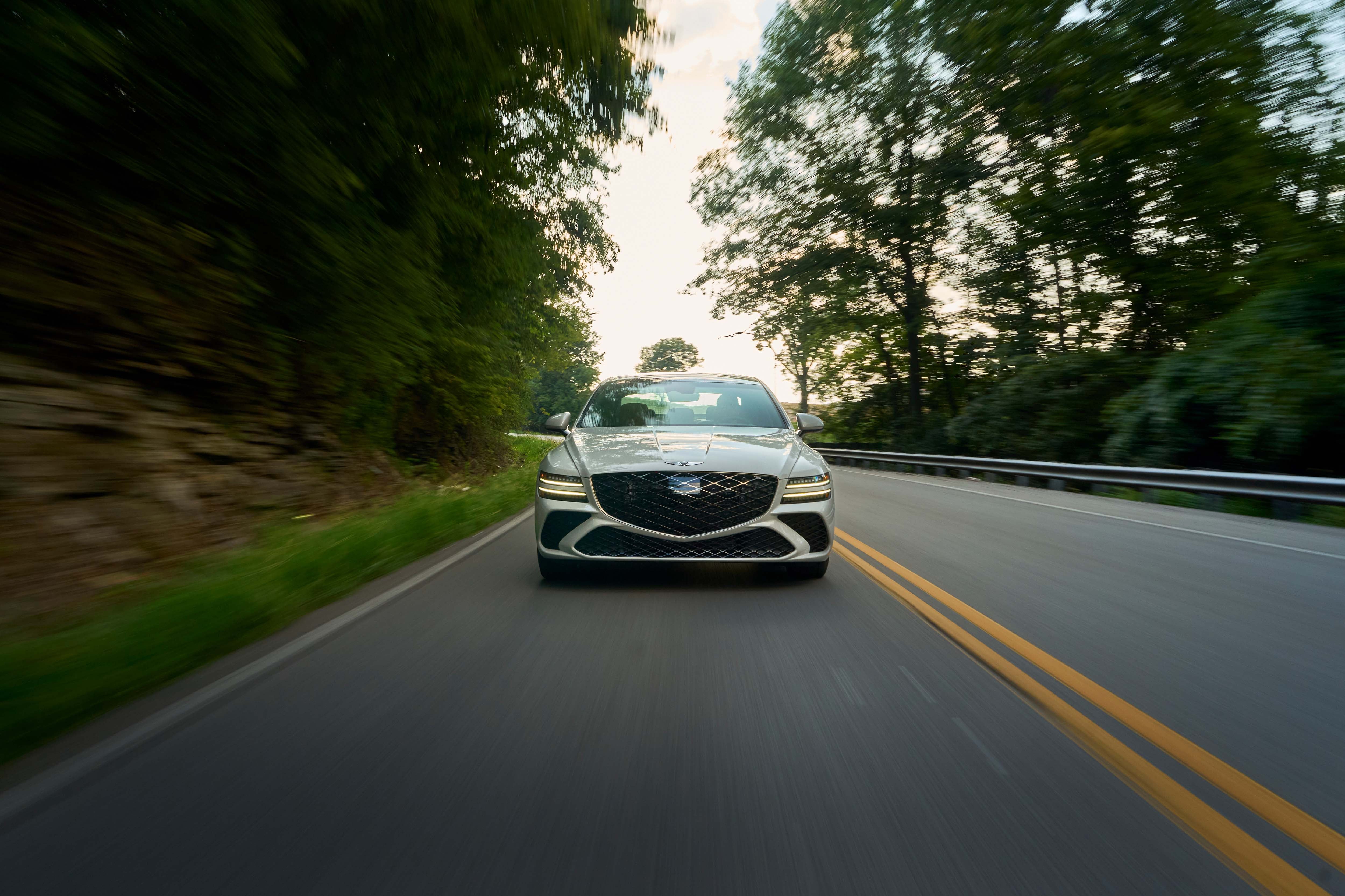 White Genesis sedan driving on a scenic two-lane road surrounded by trees at sunset.
