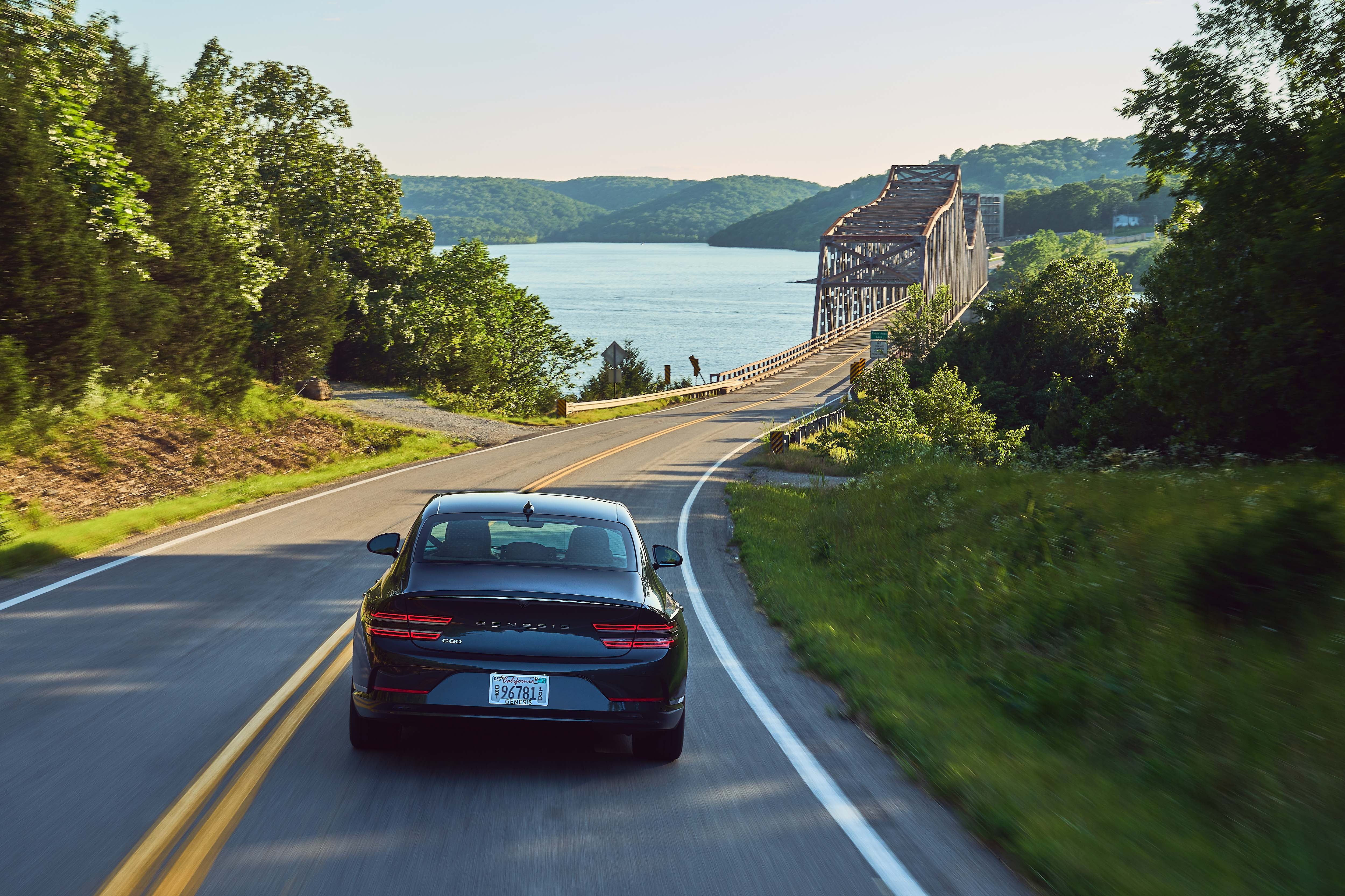 Black 2023 Genesis Electrified G80 driving towards a truss bridge over a lake, surrounded by greenery.