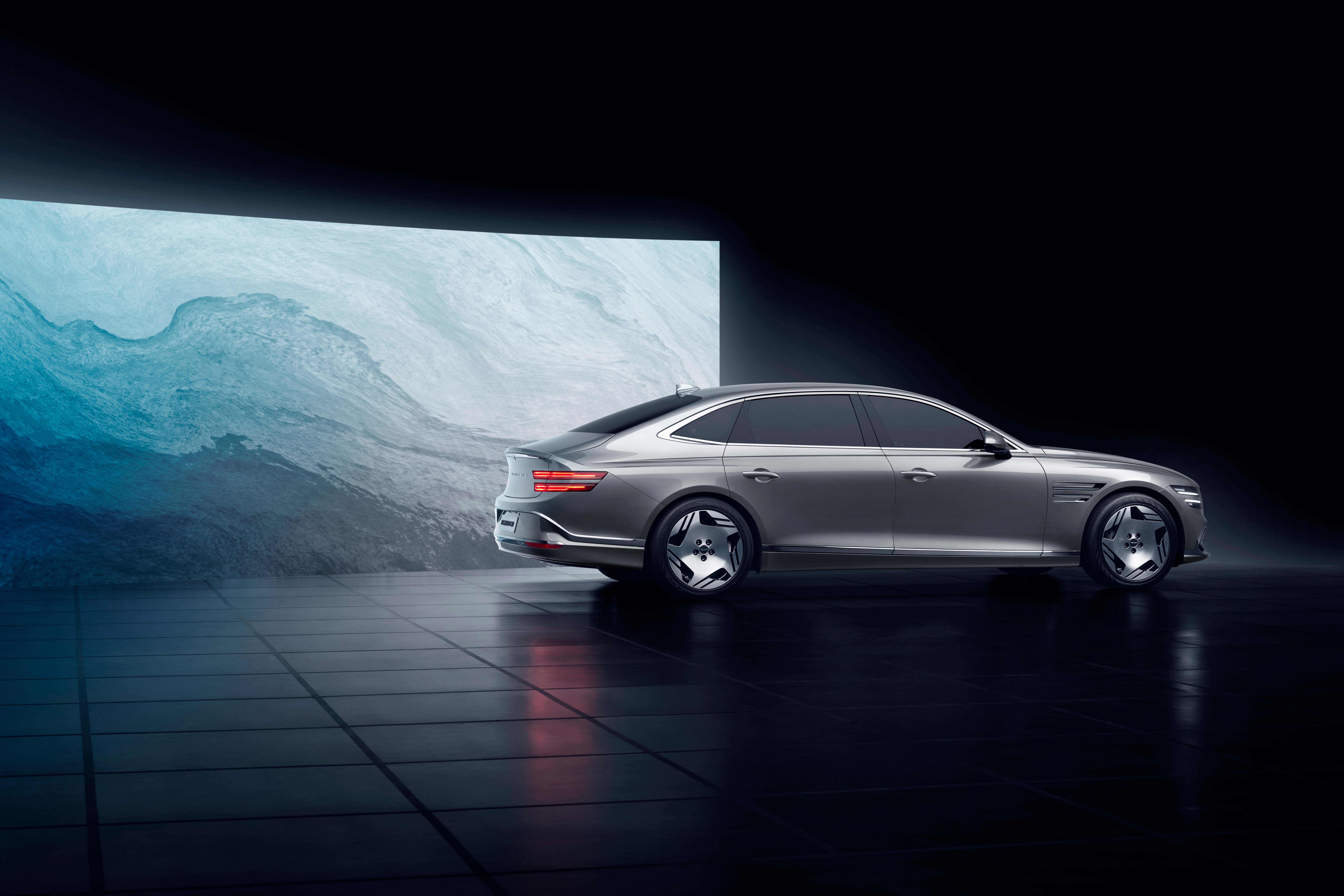 Silver 2024 Genesis Electrified G80 in a dark studio with an abstract blue and white screen backdrop.