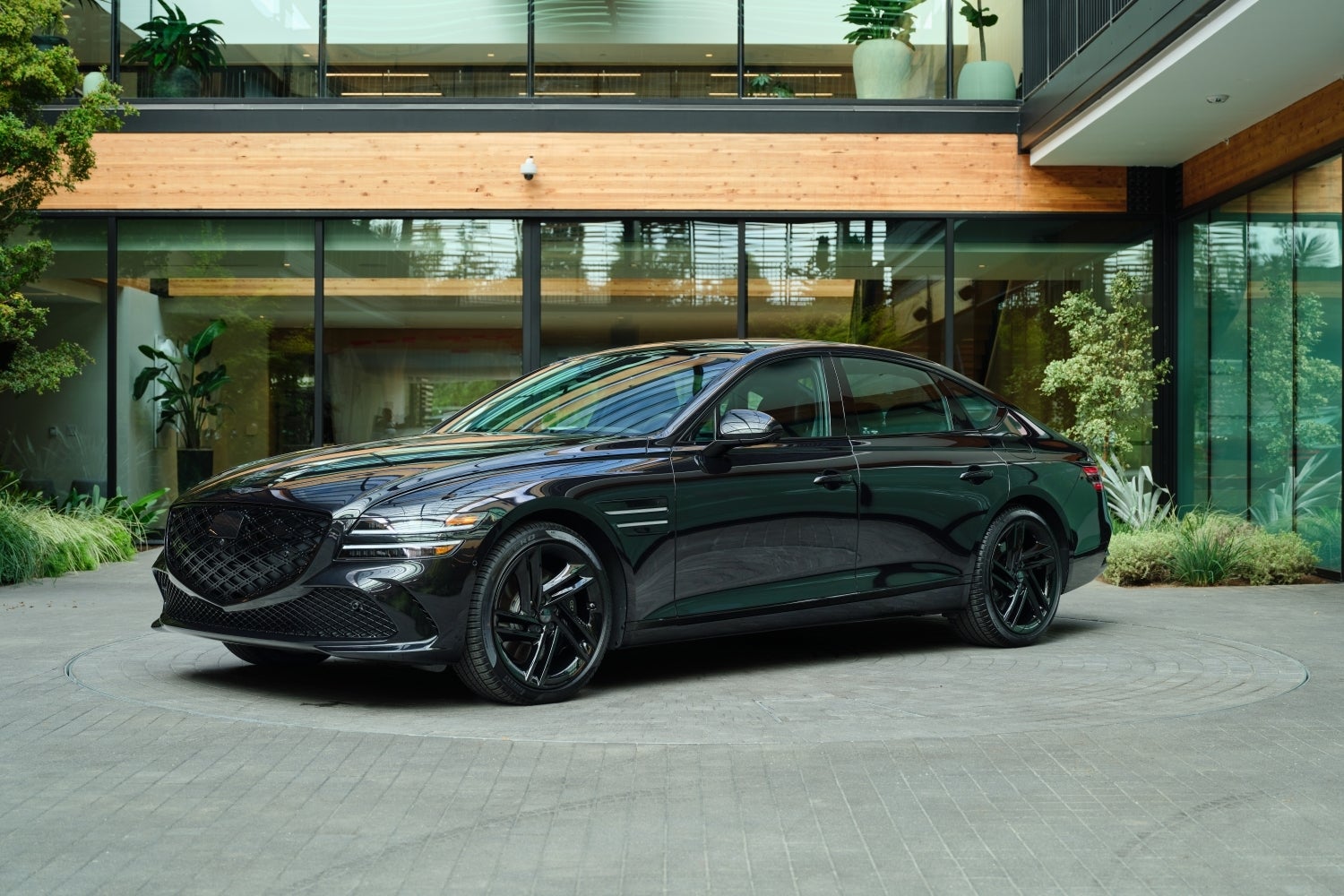 A vik black 2026 Genesis G80 is parked facing the viewer to the left on a cobblestone circle in front of a modern building made of glass and wood surrounded by plants.
