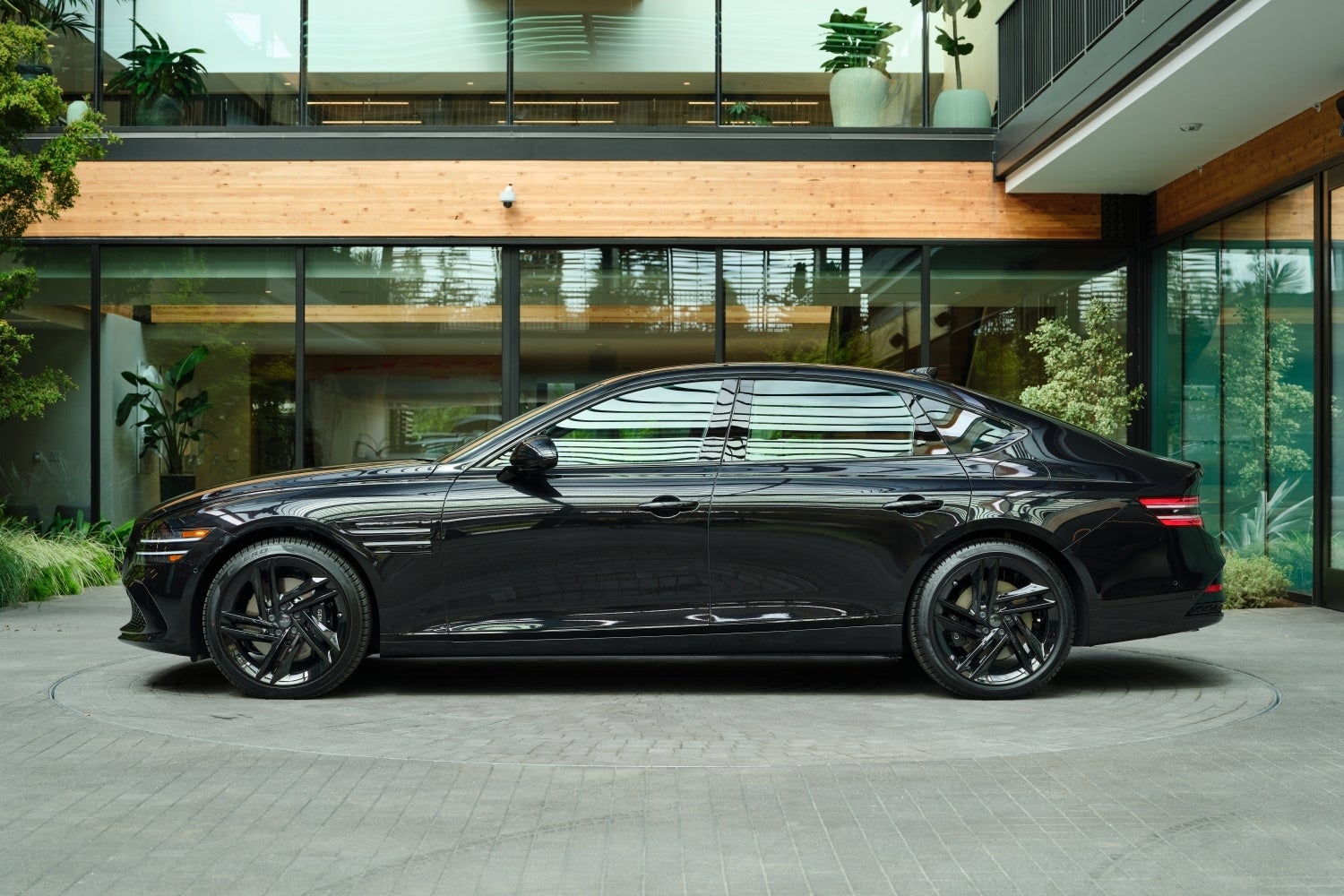 The vik black new Genesis G80 is parked facing left on a cobblestone circle in front of a modern building made of glass and wood surrounded by plants.