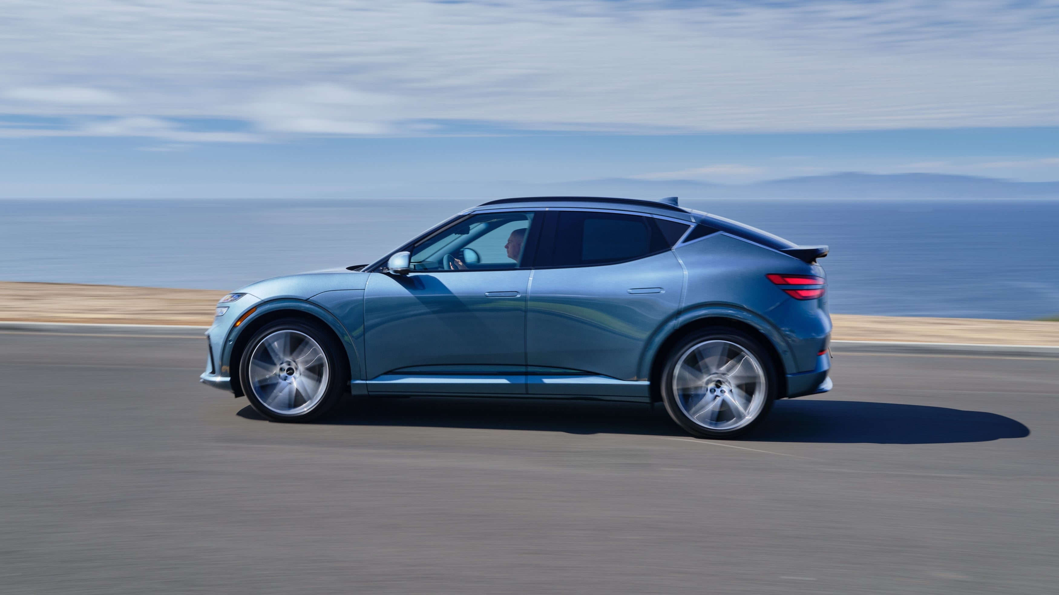 The 2026 GV60 drives to the left on a coastal road, ready for long drives in Phoenix and beyond on a single charge.