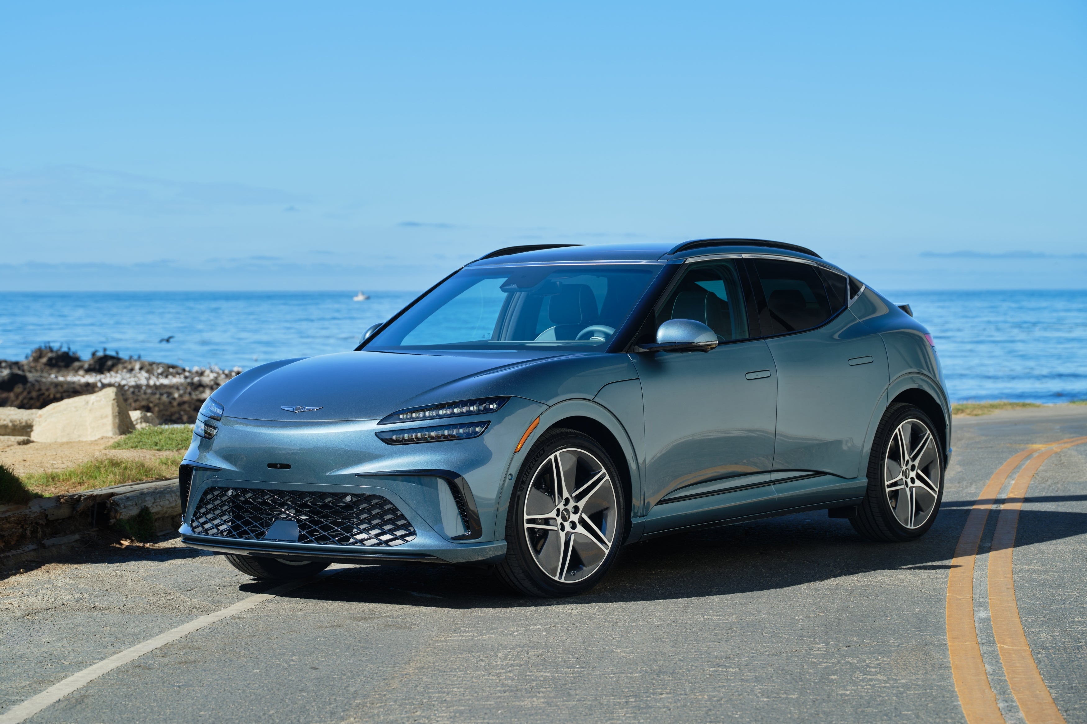 A tromso green 2026 Genesis GV60 is parked at an angle on a seaside road with the coast in the background.