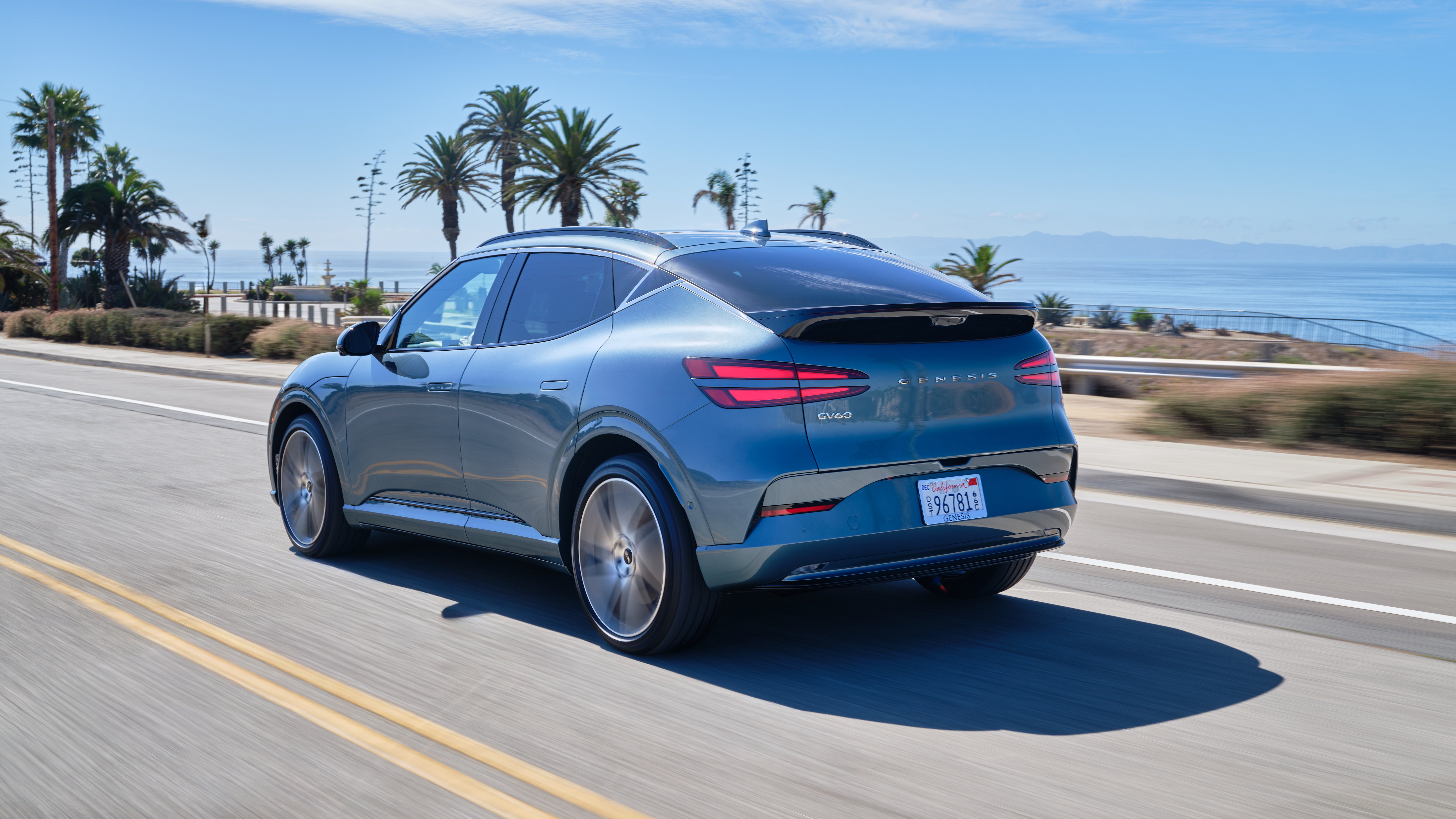 The 2026 GV60 in Phoenix is driving away the viewer to the left with many palm trees on a beachy background, with plenty of space to carry all of your adventure supplies.