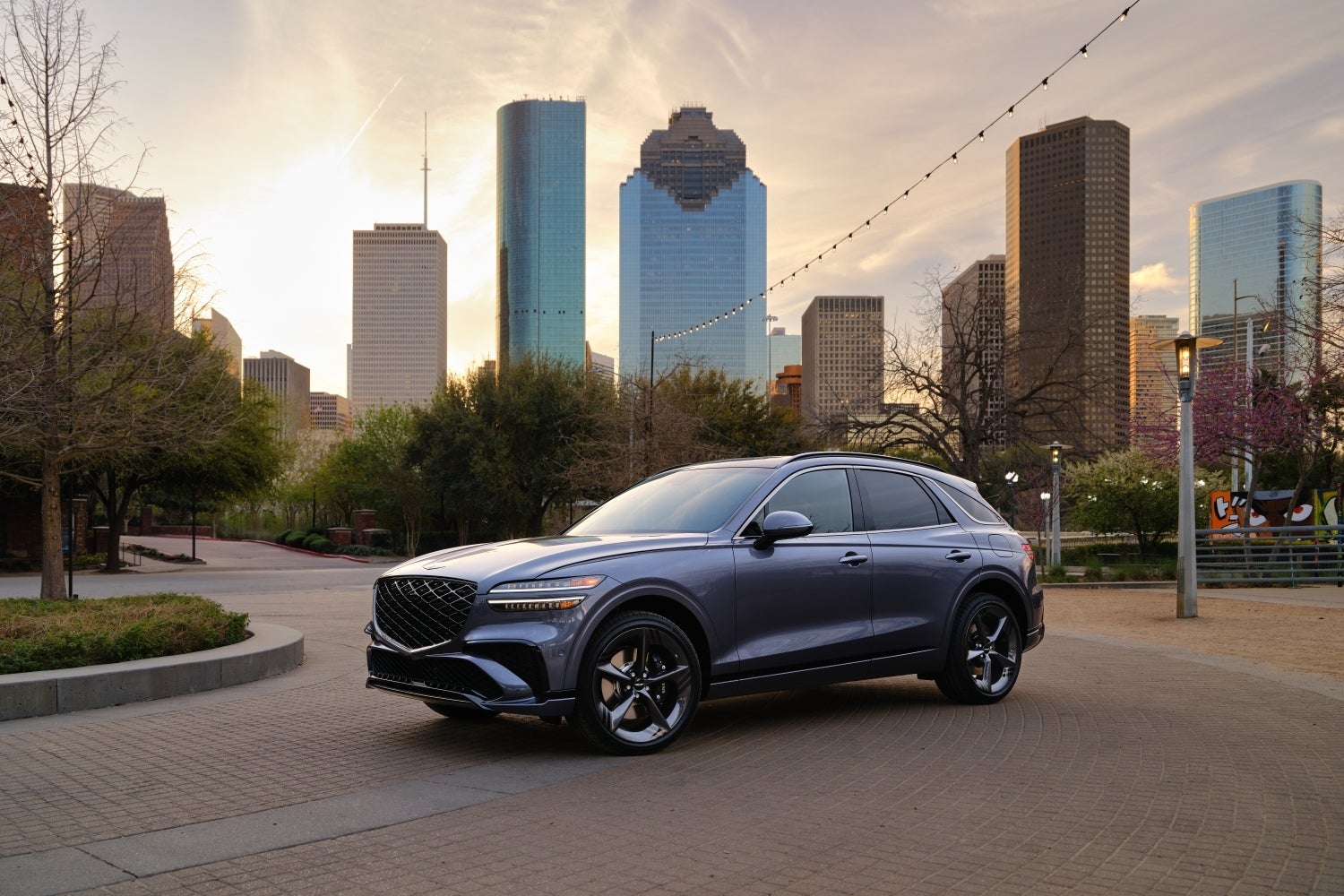The ceres blue 2026 Genesis GV70 is parked angled toward the viewer to the left on a cobblestone city street with roadside trees in the immediate background and a city skyline further back.