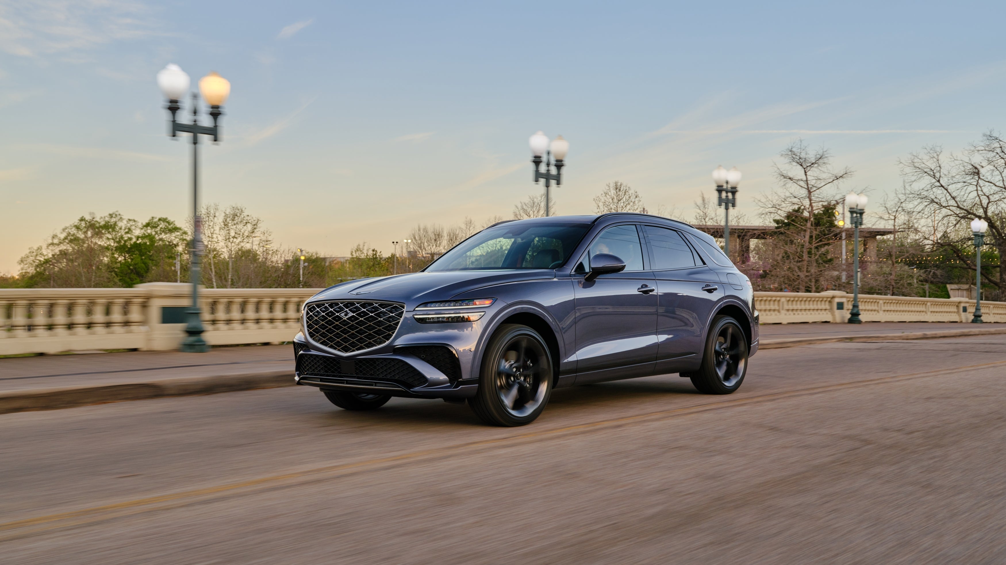A blue 2026 Genesis GV70 in Phoenix drives towards the viewer to the left on a bridge over a river in a small town.