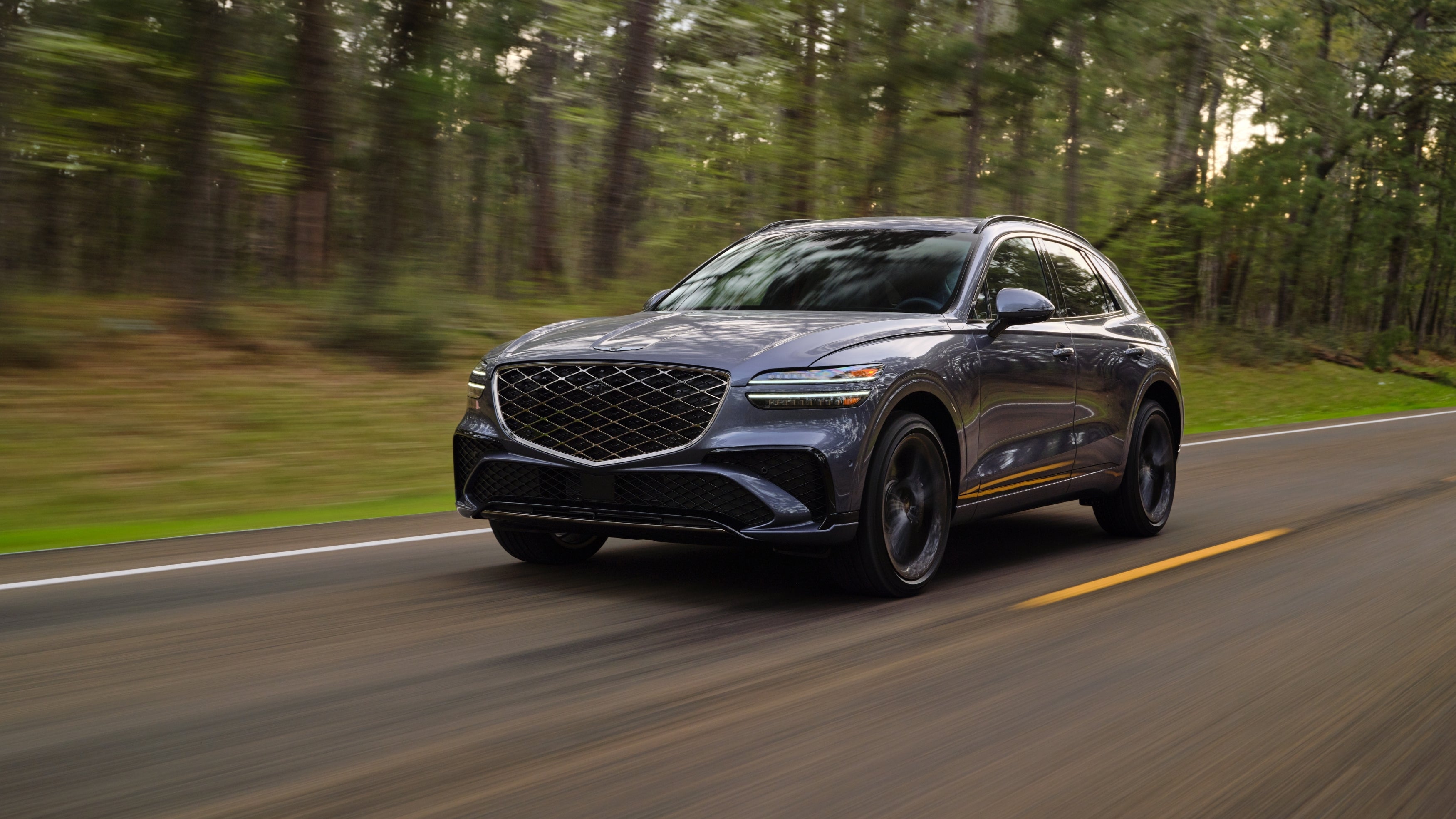 The new Genesis SUV is driving towards the viewer to the left from a low angle on a two-lane road with a forest in the background.