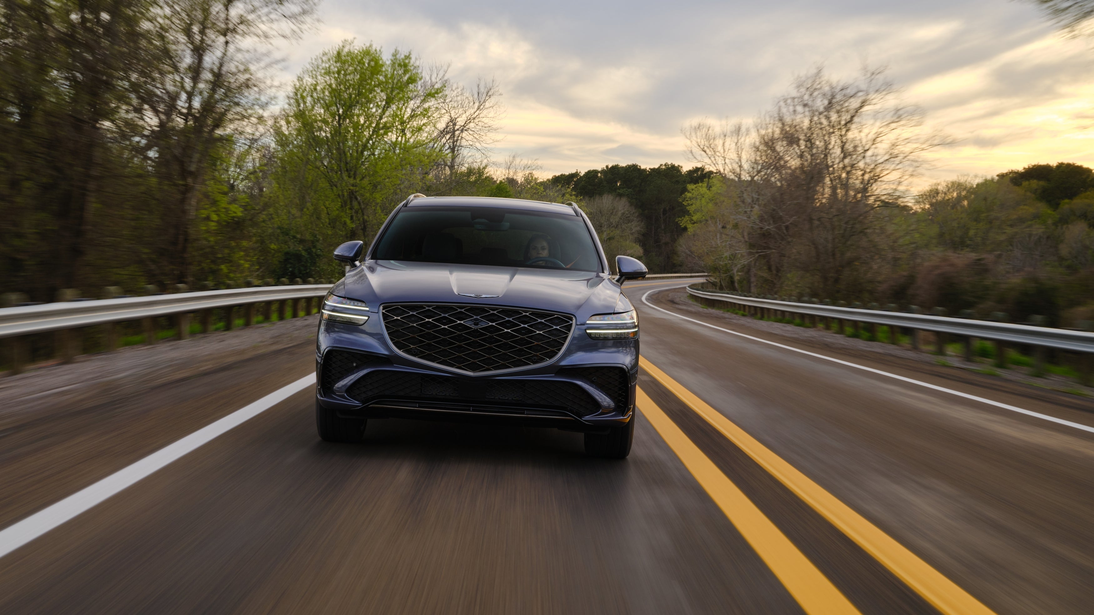 The 2026 GV70 is driving directly toward the viewer on a tilted two-lane road through trees.