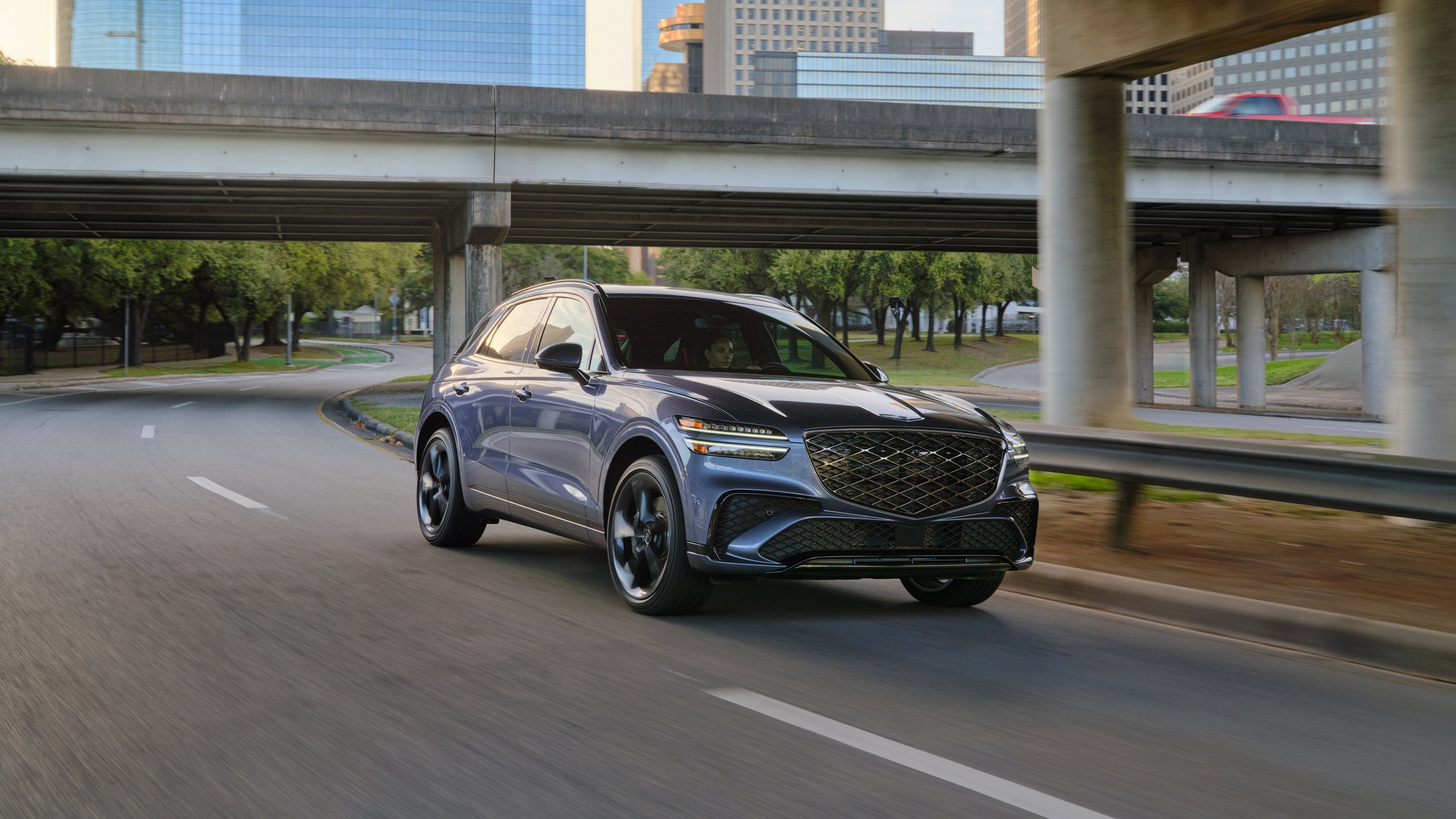 The new GV70 in Scottsdale drives toward the viewer to the right on a winding city road under an overpass.