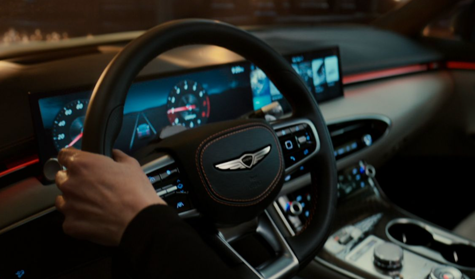 The image shows the interior of a Genesis vehicle from the driver's perspective, with a person's hands holding the steering wheel. The steering wheel is black with red stitching and features the Genesis winged emblem in the center. To the left, a digital instrument cluster displays a speedometer and other driving information, with red accents. To the right, a wide infotainment screen shows navigation or multimedia content. The dashboard features ambient lighting with a red glow along a trim line. The overall interior appears modern and luxurious, with a focus on digital displays and integrated controls.