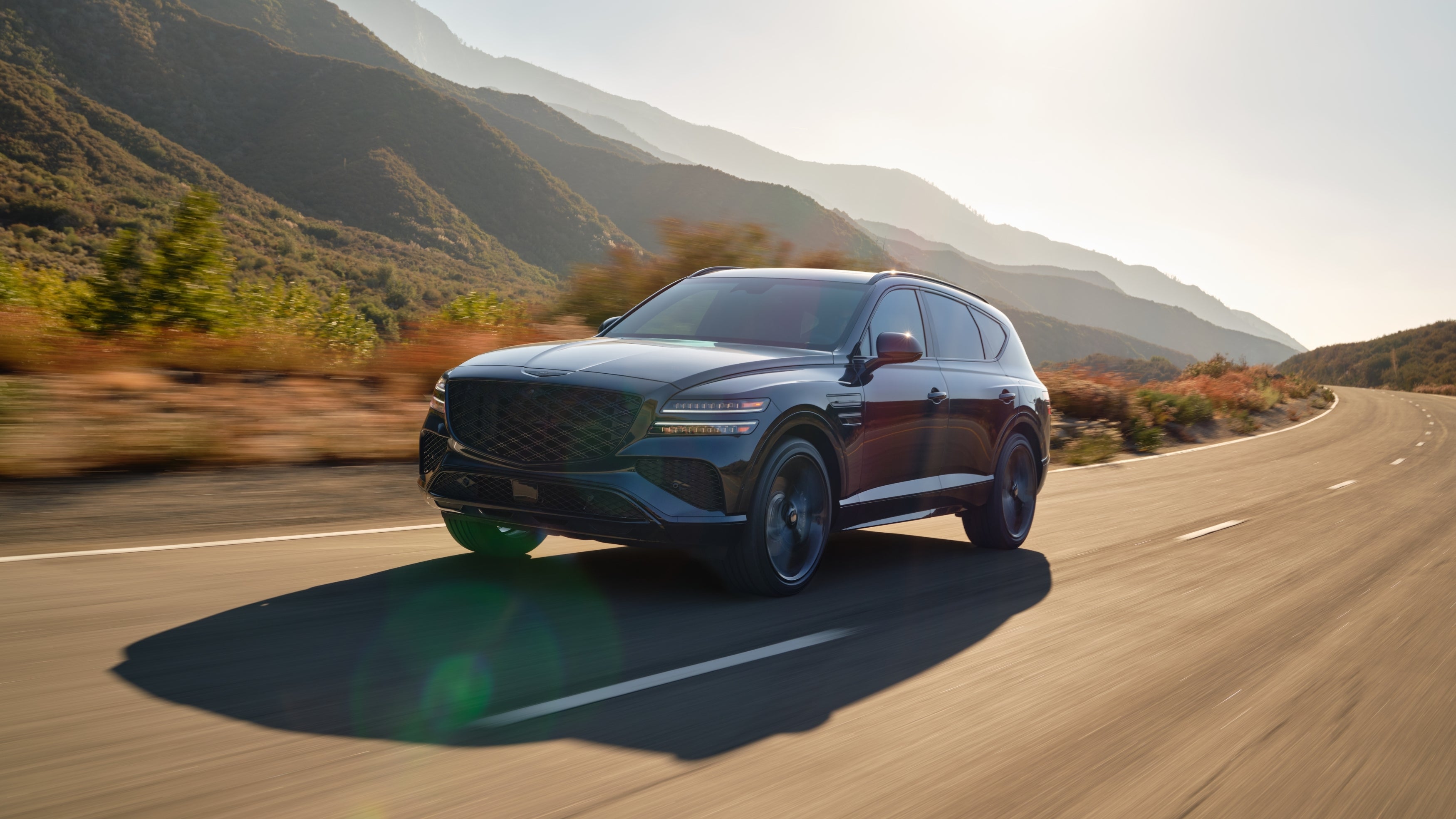 A black 2026 Genesis GV80 in Phoenix drives towards the viewer to the left on a curvy rural highway with arid mountains in the background.