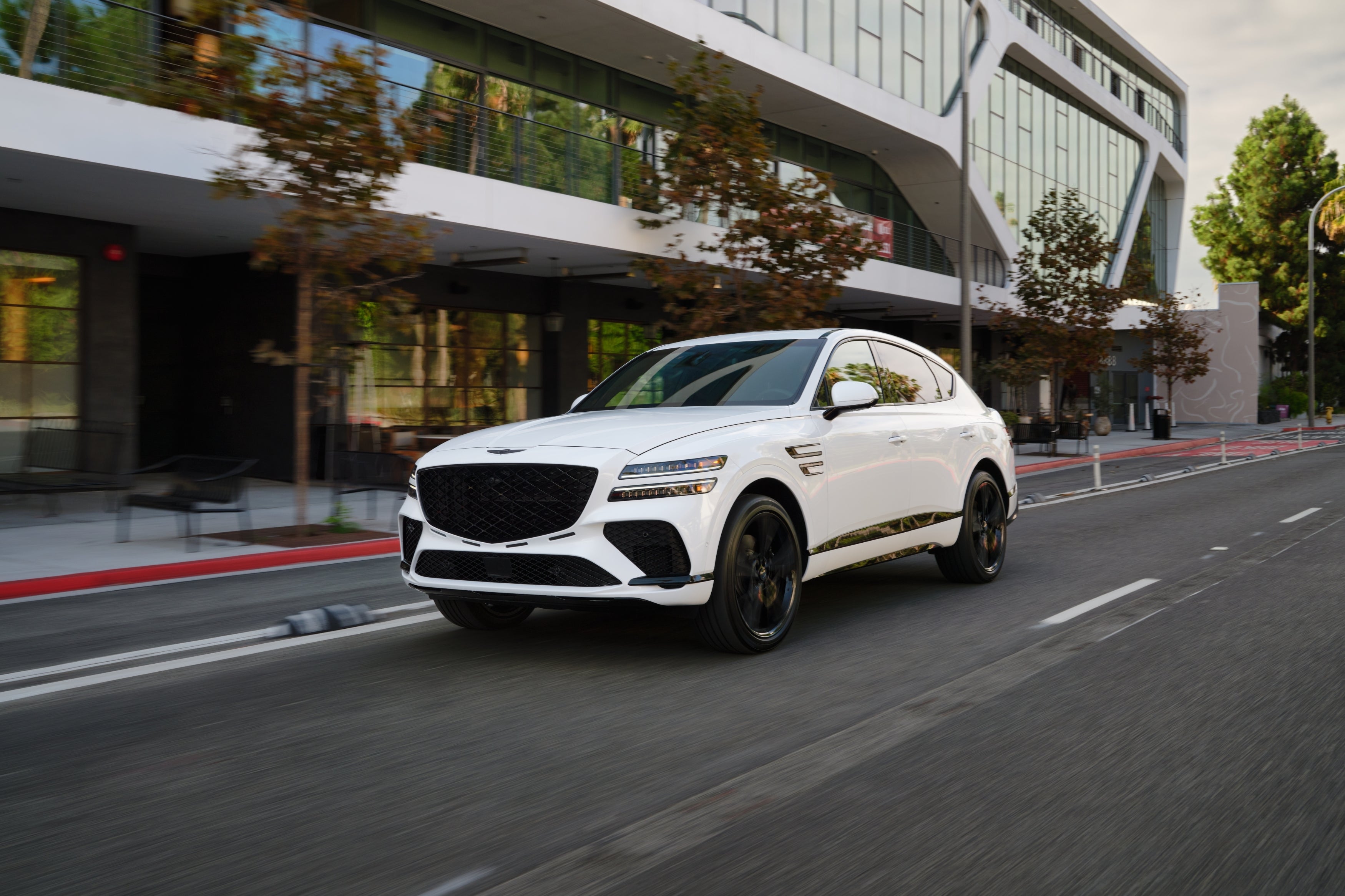 The 2026 Genesis SUV drives towards the viewer to the left on a city street in front of a large modern building.