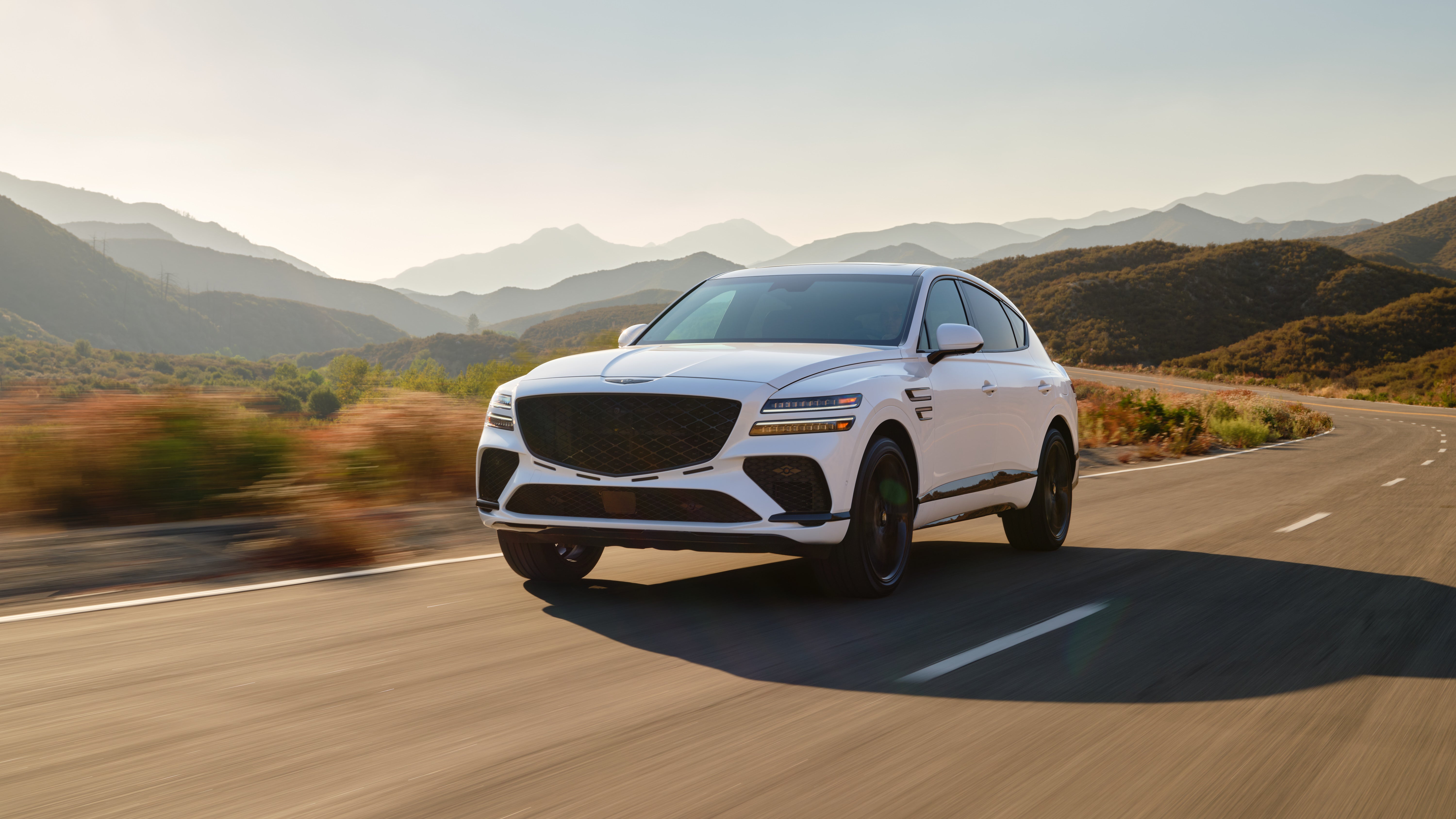 A new 2026 GV80 Coupe is driving on a winding road toward the viewer to the left with arid mountains in the background.
