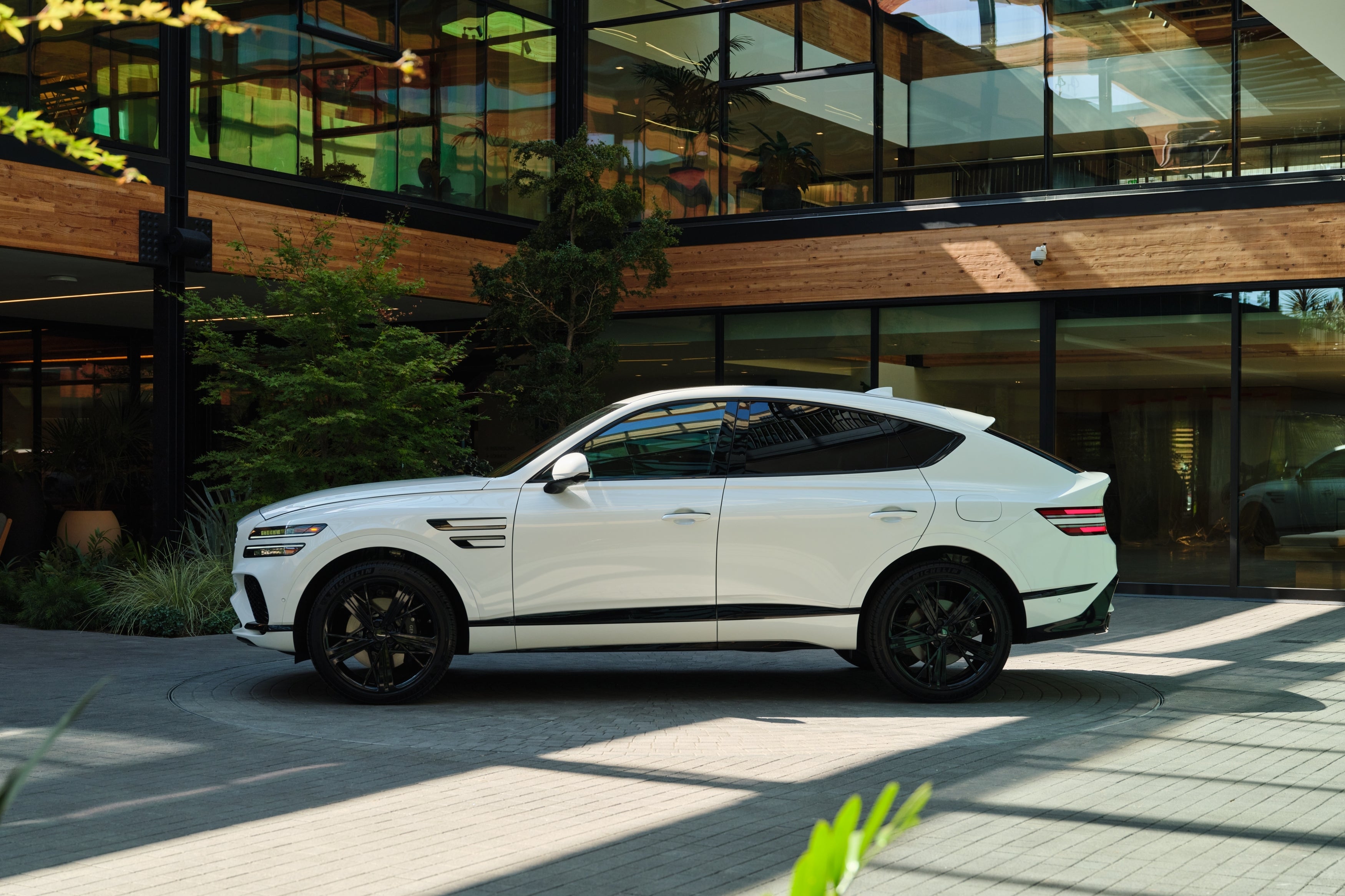 A side view of the new Genesis GV80 Coupe facing left, parked in front of a modern building surrounded by plants.