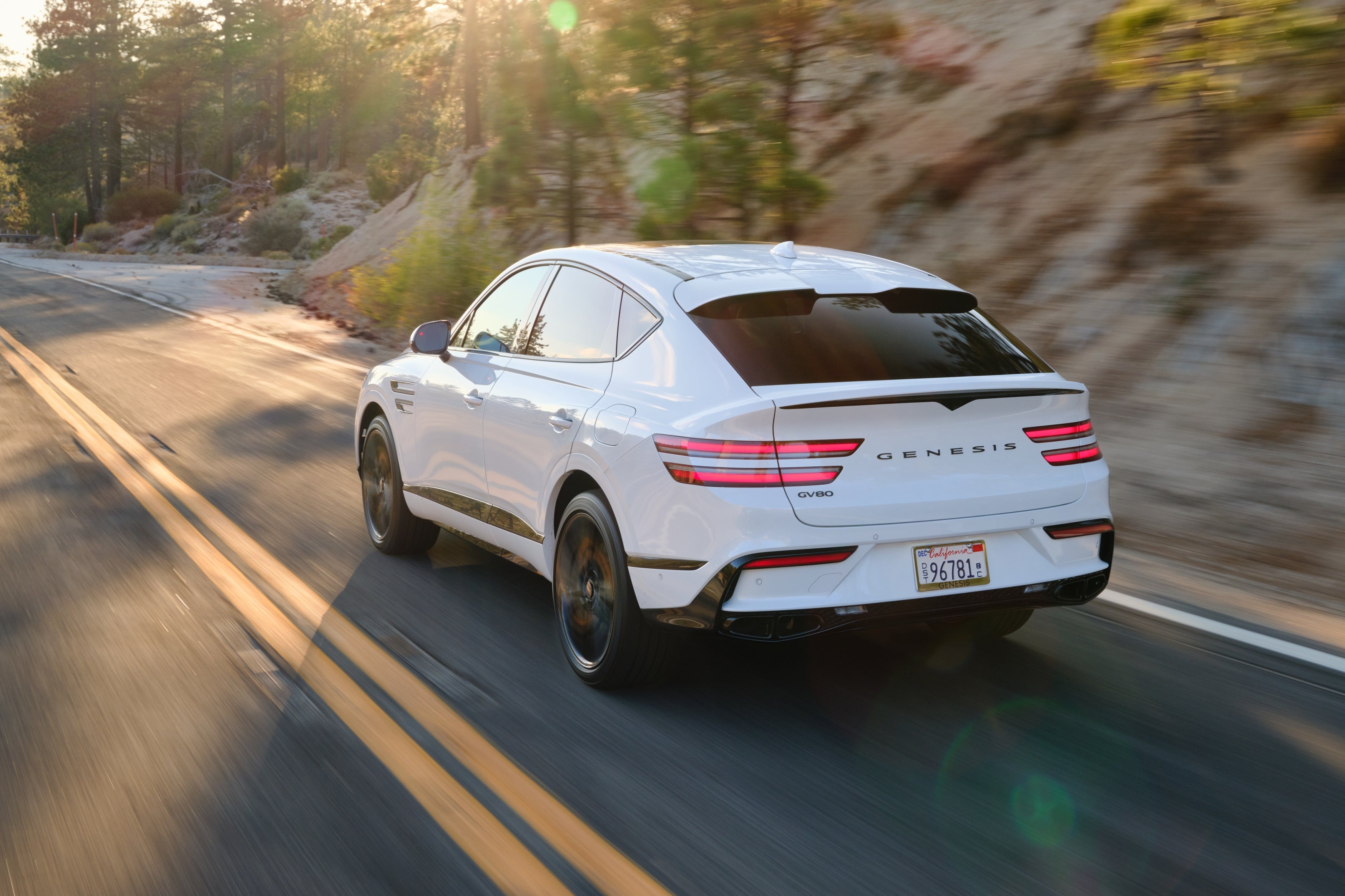 The Genesis GV80 Coupe is driving away from the viewer in Scottsdale to the left on a mountain road through young evergreen trees.