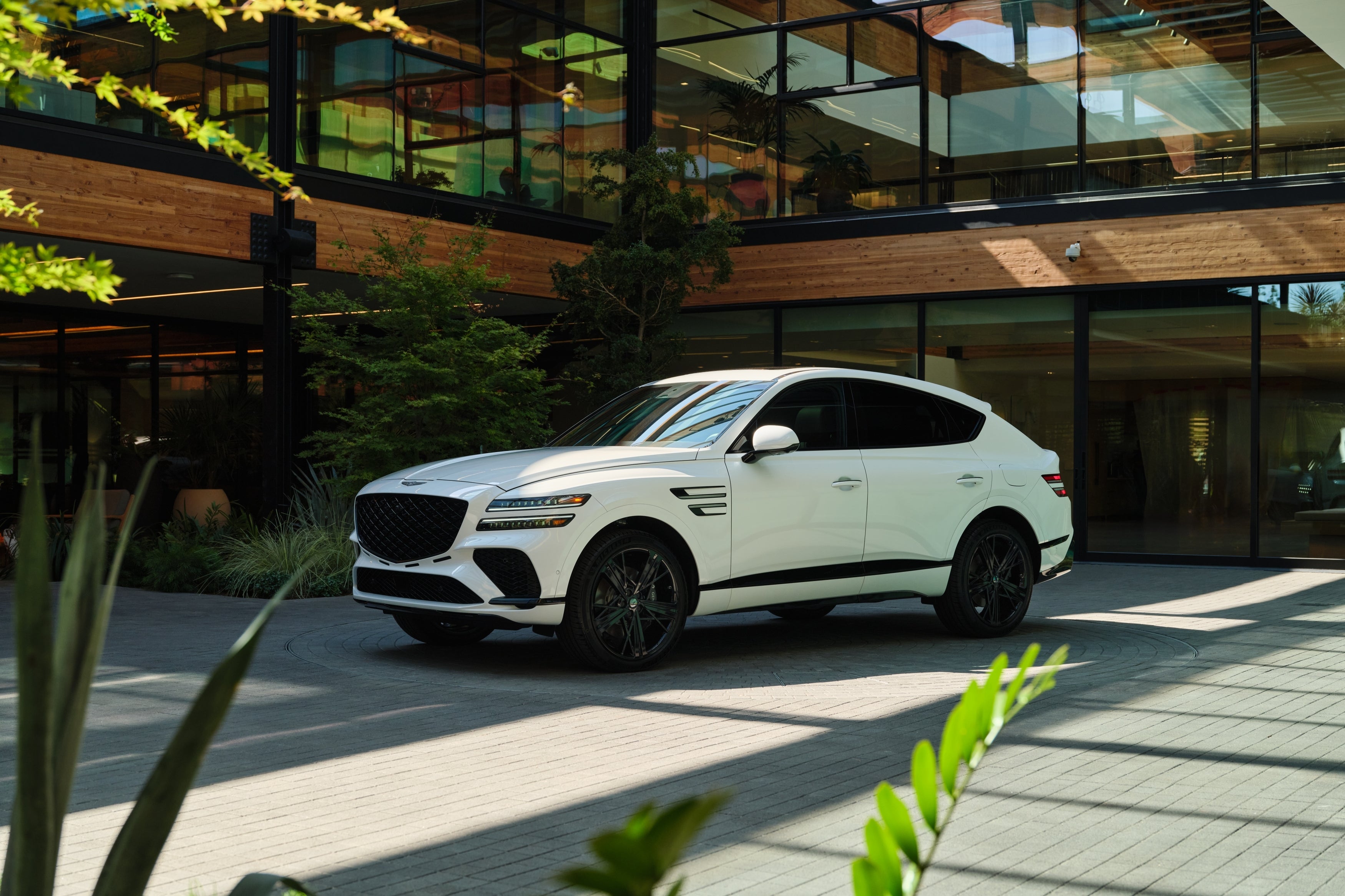 The uyuni white 2026 Genesis GV80 Coupe is parked facing the viewer to the left in front of a modern building made of wood and glass and surrounded by plants.