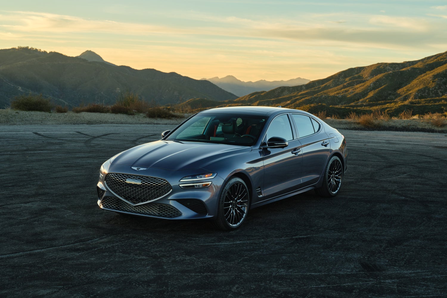 A Prestige Graphite 2026 Genesis G70 in Scottsdale is parked facing the viewer to the left on a gravel lot in the mountains at sunset.
