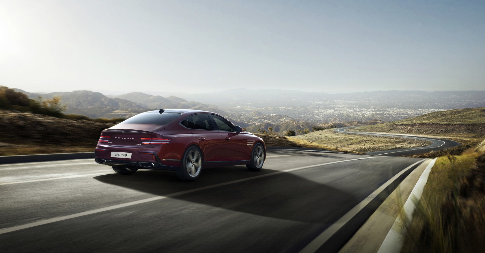 A red 2026 Genesis G80 in Phoenix drives away from the viewer to the right on a rural two-lane road passing through rolling hills.