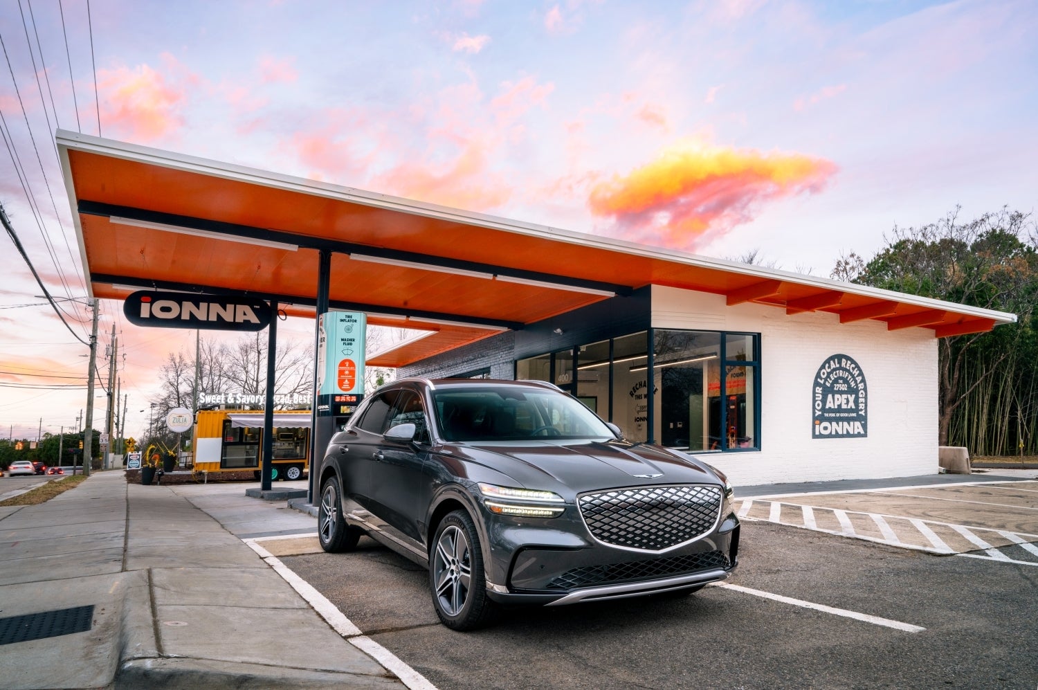 A savile silver 2026 eGV70 drives toward the viewer slightly to the right, exiting a retro gas station that has been converted into an electric charging station.