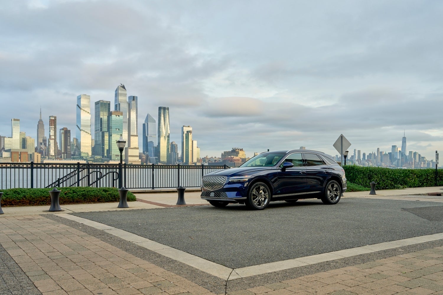 A ceres blue Genesis Electrified GV70 is seen at a distance at three quarters view parked on a paved patio with a river and a city skyline in the background