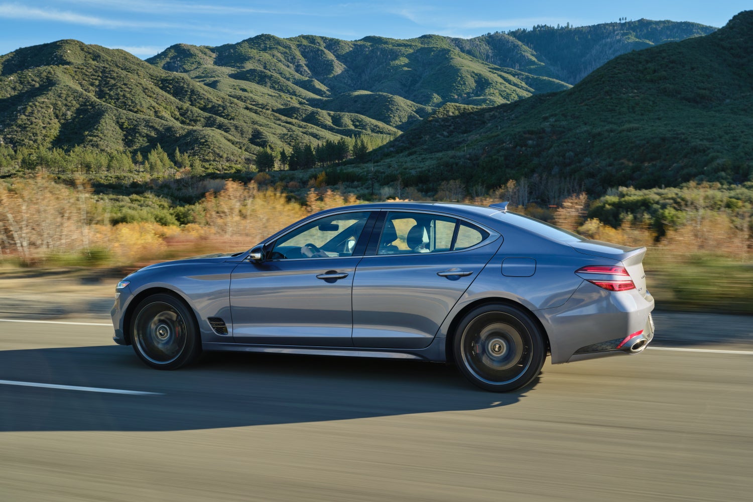 A savile silver new G70 drives to the left on a rural road with a lush green mountain in the distance.