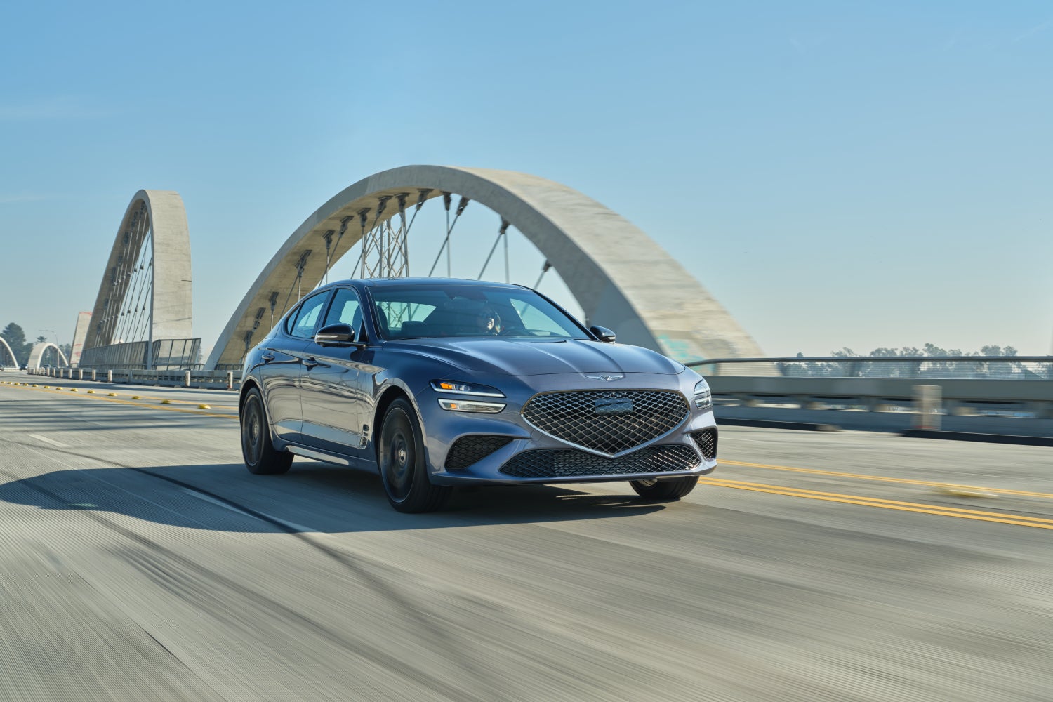 A savile silver new Genesis sedan drives toward the viewer to the right on a wide bridge over a river on a sunny day in Phoenix.