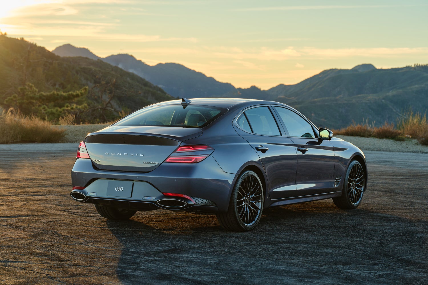A grey 2026 Genesis G70 in Scottsdale is parked facing away from the viewer to the right on a gravel road high in the mountains.