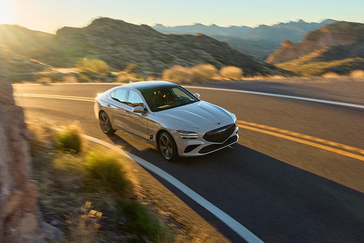 A Genesis G70 sedan driving on a winding mountain road at sunset.