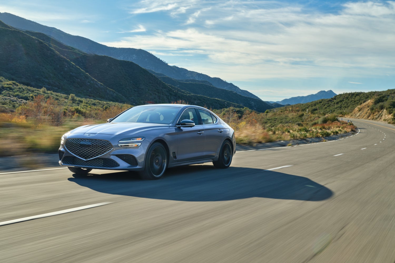 A silver 2026 Genesis G70 in Scottsdale drives towards the viewer to the left on a curvy two-lane road through an arid environment between mountains.