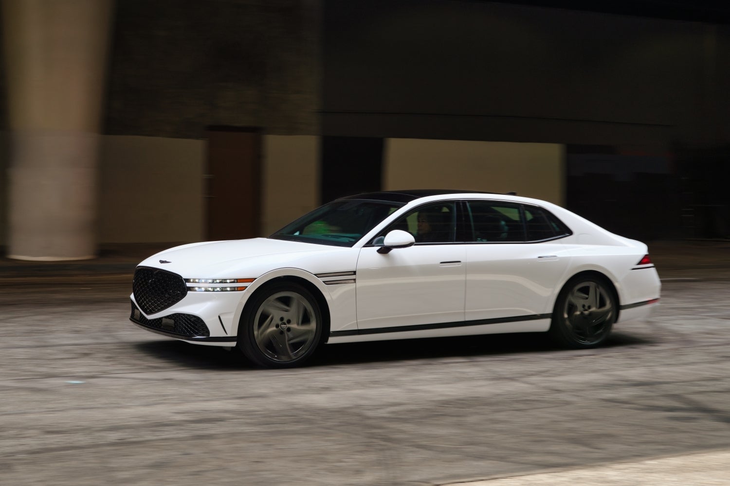 A white 2026 Genesis G90 in Phoenix drives to the left ona city street in front of a dark concrete building.