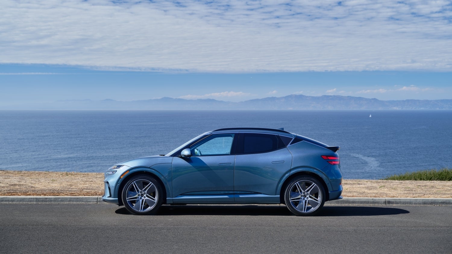 A blue 2026 Genesis GV60 in Scottsdale drives to the left on a coastal road with the sea in the background.