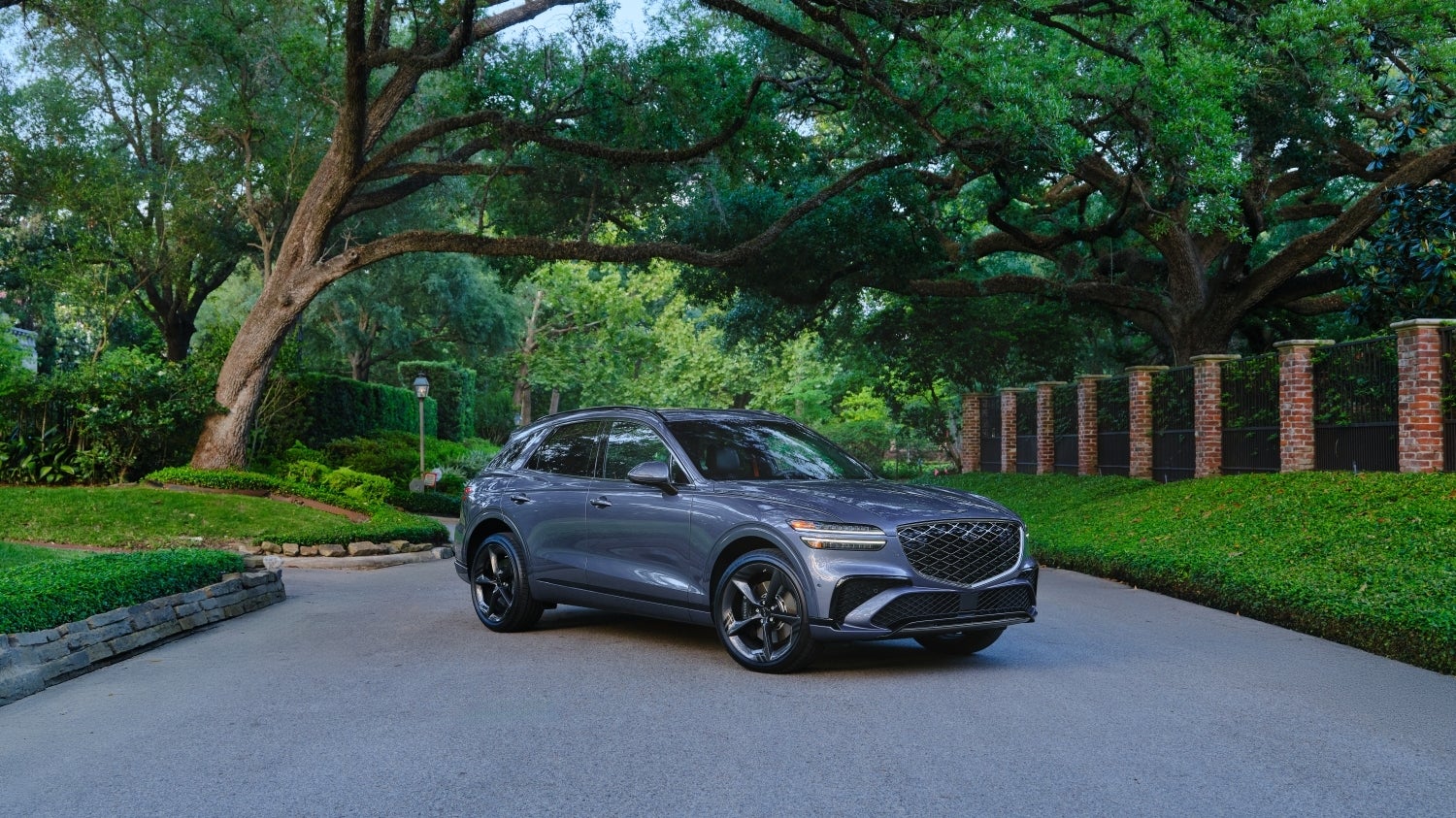 A blue 2026 Genesis GV70 in Scottsdale is parked facing the viewer to the right on an old suburban street filled with trees and greenery.