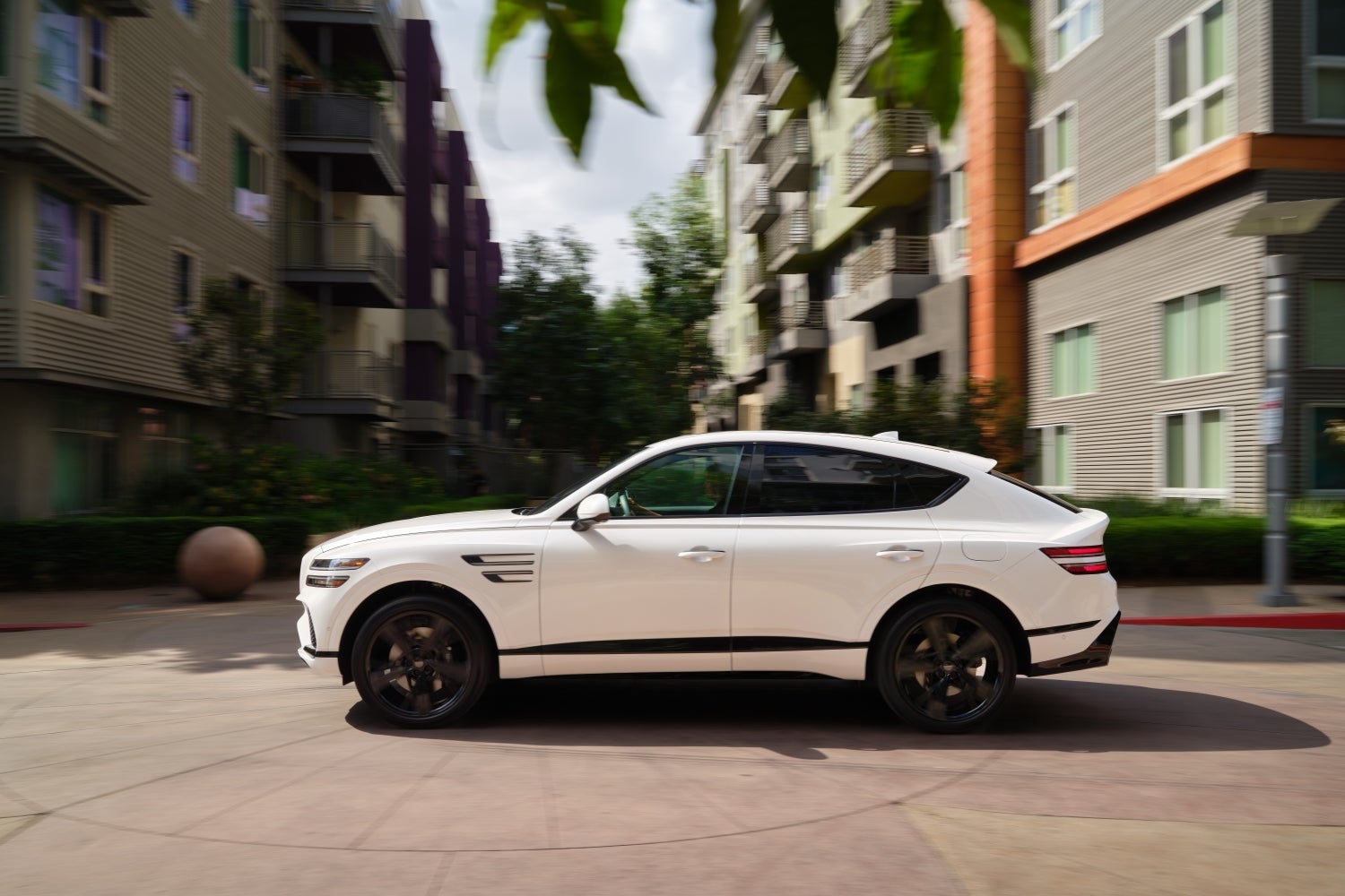 A white 2026 Genesis GV80 Coupe in Scottsdale drives to the left on a city street in front of an apartment complex.