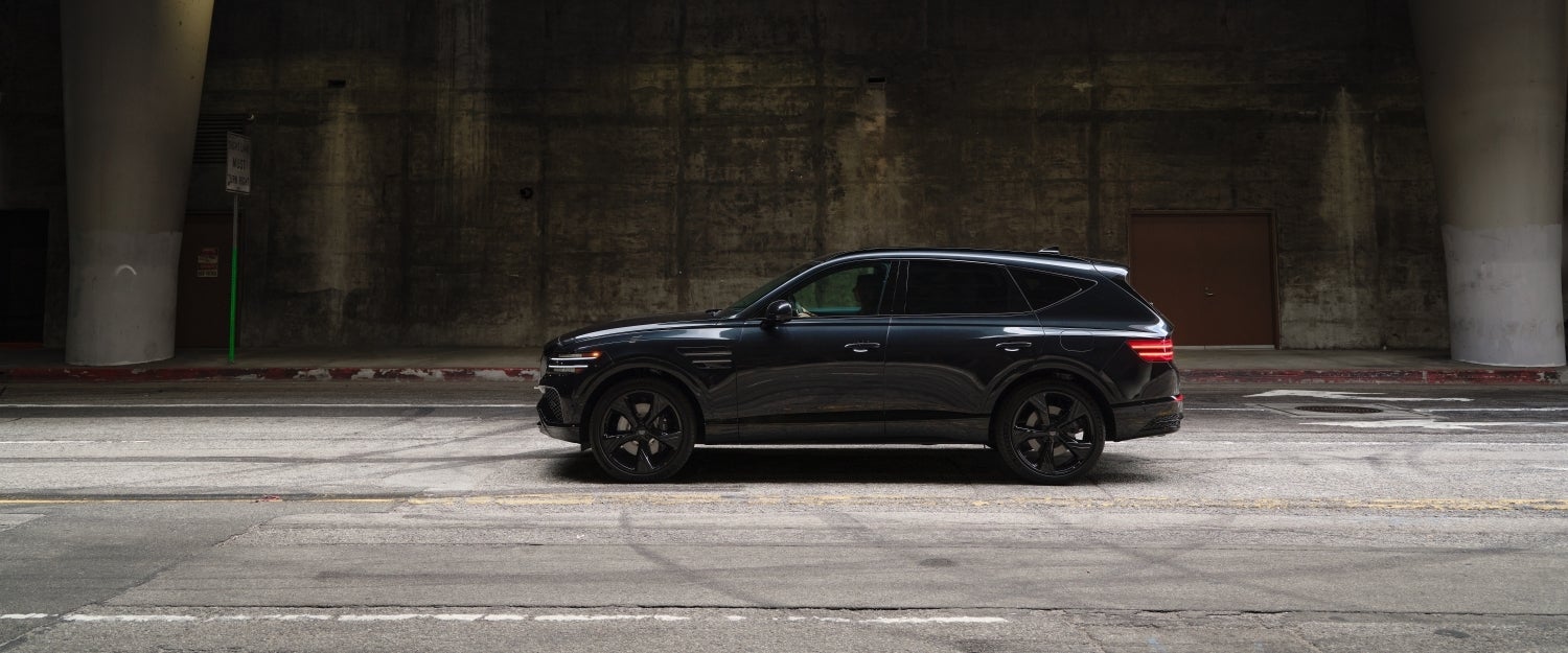 A black 2026 Genesis GV80 in Scottsdale drives to the left on a city street in front of a dark concrete building.