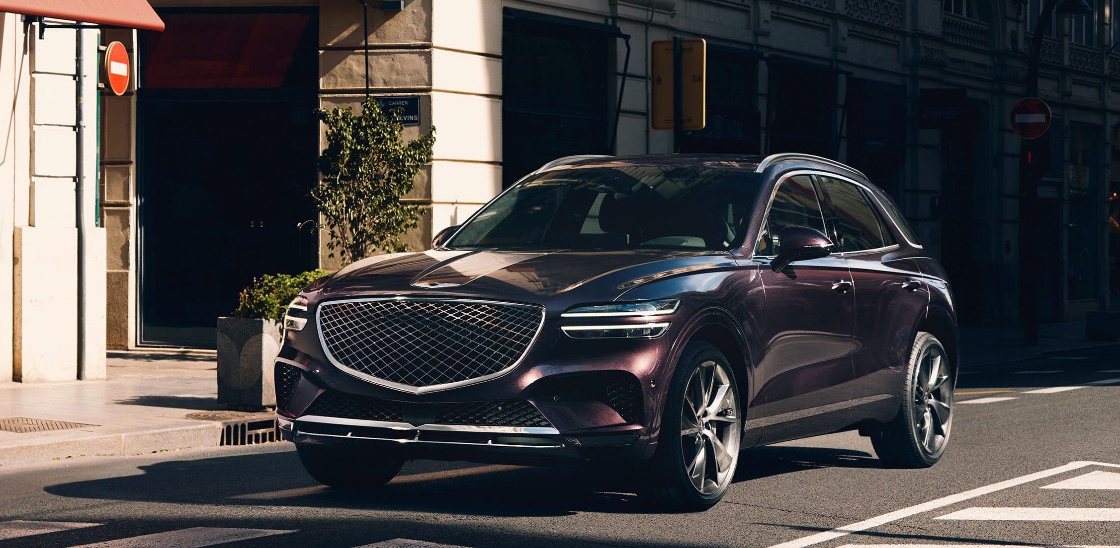 A dark red Genesis GV70 driving on a city street.
